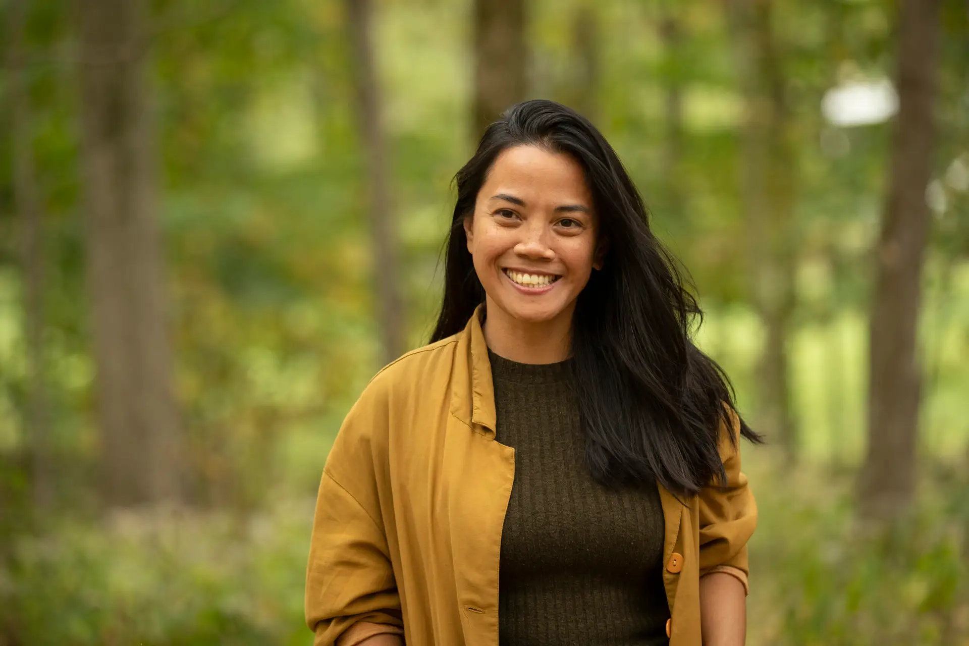 A headshot of a woman with long dark hair. She is smiling and wearing a tan shirt over a ribbed brown sweater. The background includes out of focus tree trunks and leaves.