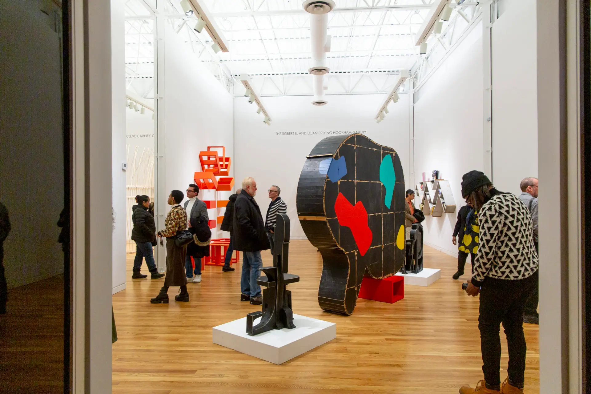 Crowd of people surround large designed objects in white open gallery.