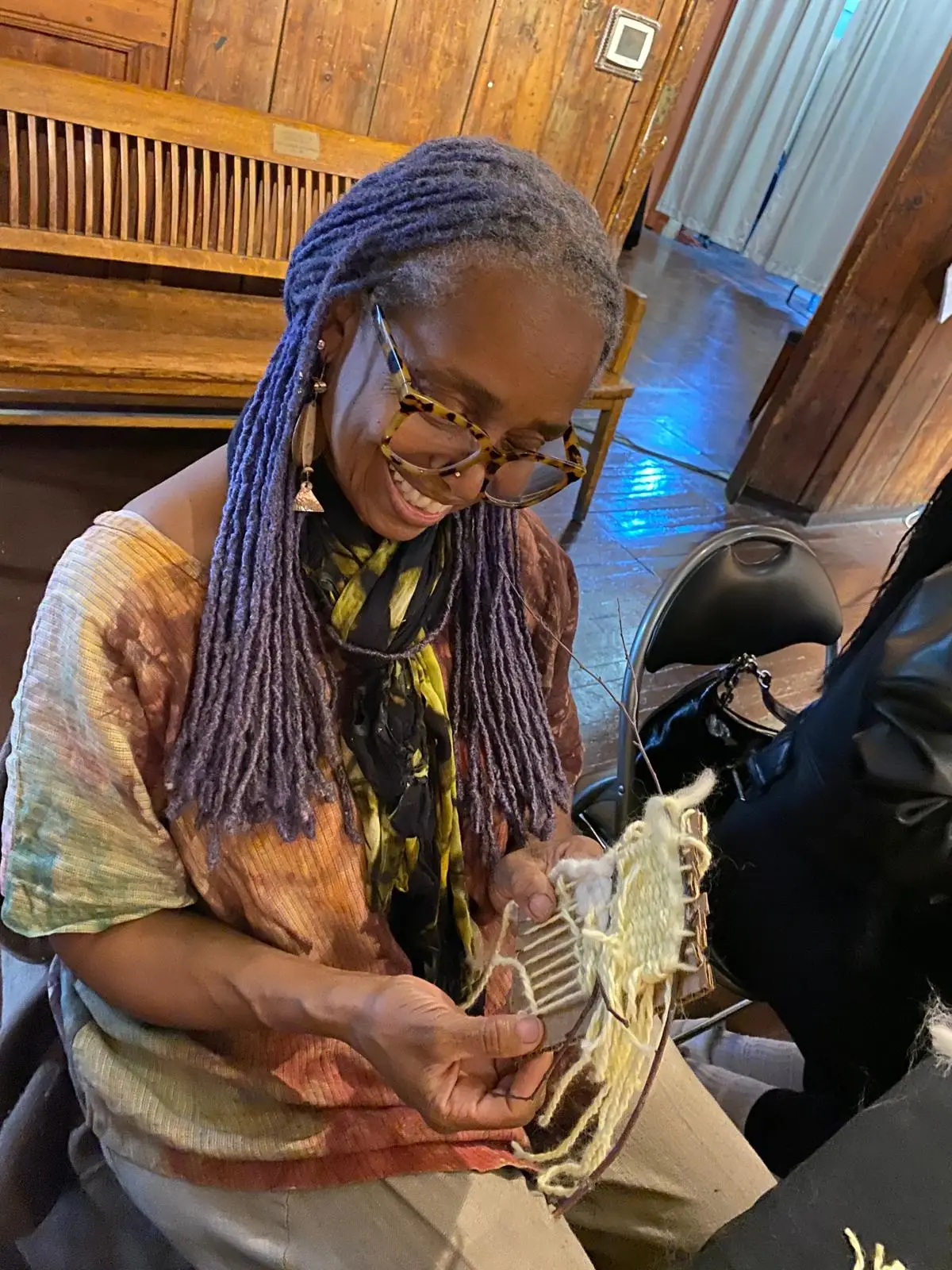 A woman with long hair and glasses is seated while working on a hand-held weaving project.